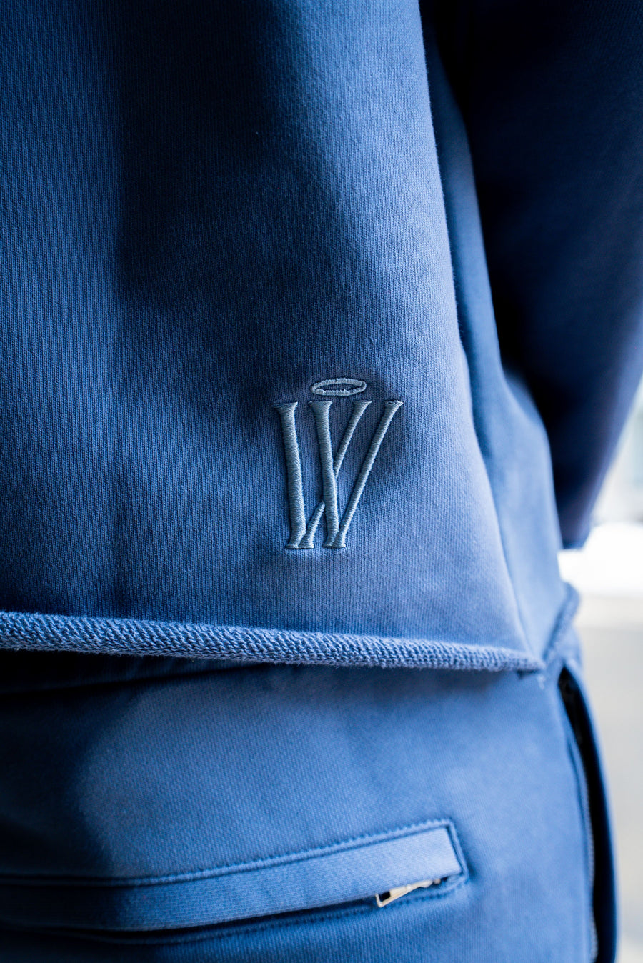 A close-up of a blue textured fabric garment featuring an embroidered stylized 'W' logo with a halo on the chest and a zippered pocket on the lower section.